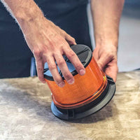 Person holding an orange and black Feniex AM600 Beacon on a wooden surface